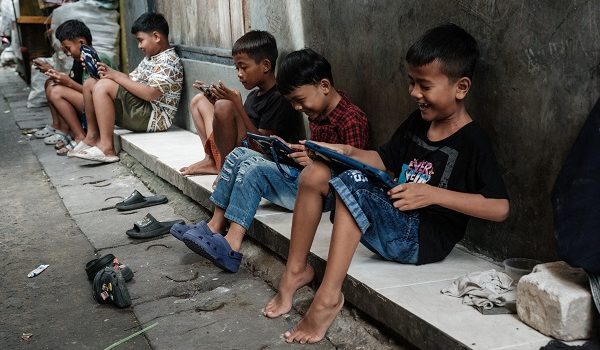 Children play online games against each other on smartphones and tablets along an alley in Jakarta on March 26, 2026. YouTube, TikTok, Facebook, Instagram, Threads, X, Bigo Live and Roblox -- deemed "high-risk" platforms by authorities in the Southeast Asian nation -- will from March 28, 2026, start deactivating underage accounts. (Photo by YASUYOSHI CHIBA / AFP) / TO GO WITH AFP STORY: Indonesia-culture-Internet-law, FOCUS by Marchio GORBIANO