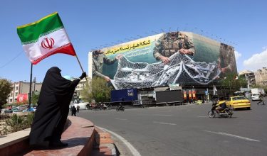 A woman holds Irans national flag while standing near a billboard with a sentence reading The Strait of Hormuz remains closed at the Enqelab Square in Tehran, on April 5, 2026. Oman and Iran held talks on easing passage through the Strait of Hormuz, the Omani state news agency reported on April 5, with the key shipping chokepoint effectively closed due to war in the Middle East. (Photo by AFP) /